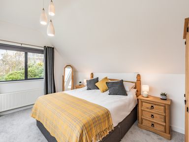 A bedroom with a bed covered with a yellow throw and grey and yellow pillows bedside tables with lamps a tall mirror and a window with dark curtains at Beach View in Combe Martin