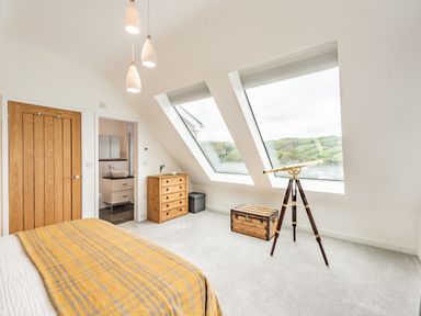 A bedroom with a bed covered by a yellow blanket chest of drawers a telescope and large slanted windows showing a view of green hills at Beach View in Combe Martin