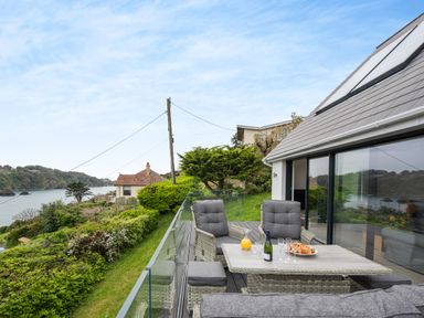 A patio with grey cushioned chairs and a table set with drinks and food overlooking water and greenery at Beach View in Combe Martin