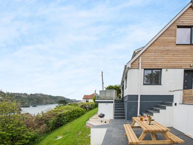 A patio with a wooden picnic table and a charcoal grill next to a house with wooden siding near a grassy hill overlooking water at Beach View in Combe Martin