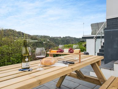 An outdoor wooden picnic table with a bottle of wine glass of red wine bread cheese platter and grapes on a terrace with a view of green hills at Beach View in Combe Martin