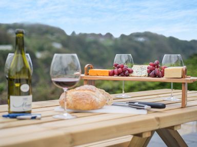 A wooden picnic table with a loaf of bread knives bottle of wine glasses cheese and grapes outdoors at Beach View in Combe Martin