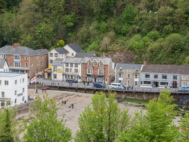 A row of buildings along a street with people walking and cars parked near a sandy area and trees at Beach View in Combe Martin