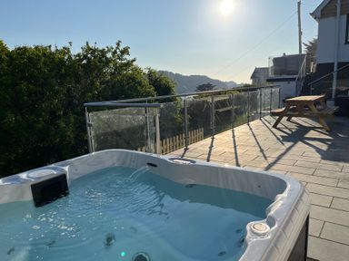 A hot tub on a tiled terrace with a wooden picnic table and glass railing overlooking trees and hills at Beach View in Combe Martin