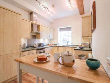 A kitchen with wooden cabinets a countertop with a stove and cooking pots a wooden island table with a cake a teapot and cups at Feathers Blythburgh in Reydon