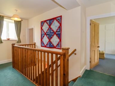A upstairs hallway with a wooden staircase railing a quilt hanging on the wall and a window with curtains at Canny Croft Chilton Polden near Street