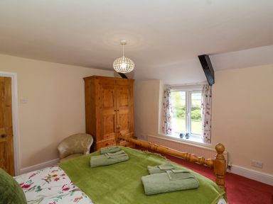 A bedroom with a wooden bed green blankets wooden wardrobe a beige armchair and a window with floral curtains at Canny Croft in Chilton Polden near Street