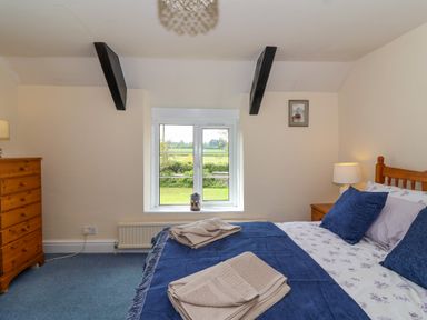 A bedroom with a bed with blue and white bedding a window showing greenery a wooden chest of drawers and a bedside table with a lamp at Canny Croft in Chilton Polden near Street