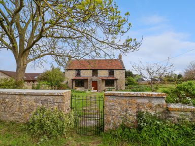 A stone house with a tiled roof behind a low stone wall and metal gate in a garden with trees at Canny Croft in Chilton Polden near Street