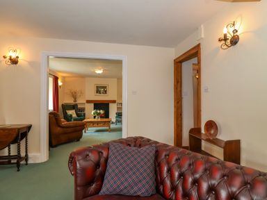 A room with a brown leather sofa and a blue plaid pillow looking into another room with green and brown armchairs a wooden coffee table and a fireplace at Canny Croft in Chilton Polden near Street
