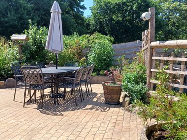 An outdoor patio with a table, six chairs, a closed umbrella, potted plants, and wooden fencing at Woodbine Cottage in Mylor Bridge Nr Falmouth