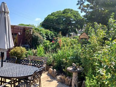 A garden with a patio table and chairs a closed umbrella plants a birdbath and a shed at Woodbine Cottage in Mylor Bridge Nr Falmouth