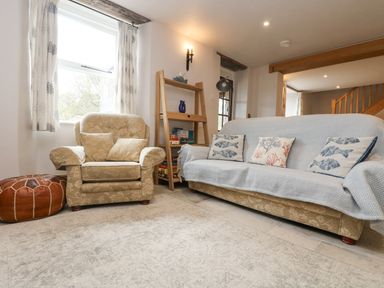 A living room with patterned armchair and sofa with cushions by a window with curtains and wooden bookshelf at Woodbine Cottage in Mylor Bridge Nr Falmouth