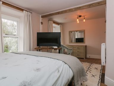 A bedroom with a bed a tv on a wooden stand a wicker chair a wooden dresser with a mirror and two windows with curtains at Woodbine Cottage in Mylor Bridge Nr Falmouth