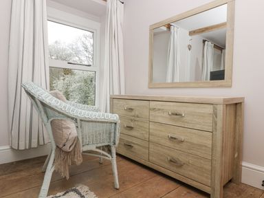 A wicker chair with a blanket next to a wooden dresser and mirror in a room at Woodbine Cottage Mylor Bridge Nr Falmouth