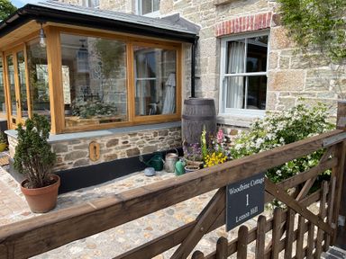 A stone cottage exterior with a wooden conservatory a water barrel plants gardening tools and a wooden gate with a sign Woodbine Cottage 1 Lemon Hill