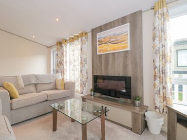 A living room with a beige sofa yellow pillows glass coffee table and wall-mounted television at Gwydn Lodge in Downton Hampshire