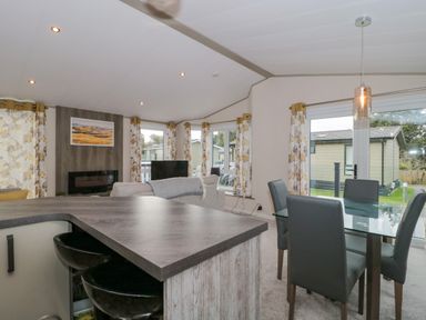 An open living and dining area with a glass dining table with chairs a kitchen counter two bar stools a gray sofa a wall-mounted fireplace multiple windows with floral curtains and a hanging light at Gwydn Lodge in Downton Hampshire