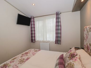 A bedroom with a bed featuring floral and plaid bedding a window with plaid curtains and a wall-mounted television at Gwydn Lodge in Downton Hampshire