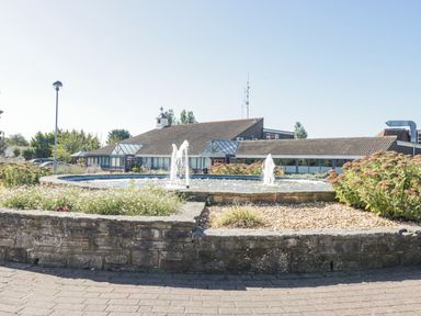 An outdoor fountain with water jets in front of a low building with sloped roofs and glass windows at Gwydn Lodge in Downton Hampshire