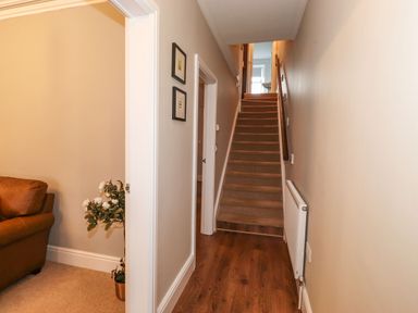 A hallway with wooden flooring leading to carpeted stairs and a partially visible room with a brown sofa and a potted plant at Snowflake Cottage in Windermere