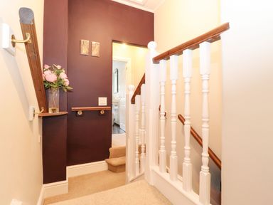 A stairway landing with wooden handrails and white balusters a purple accent wall with framed art and a vase of pink flowers at Snowflake Cottage in Windermere