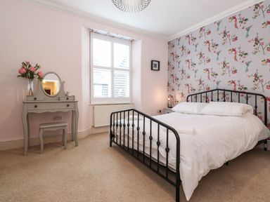 A bedroom with a metal bed frame white bedding floral wallpaper a dressing table with mirror and flowers at Snowflake Cottage in Windermere