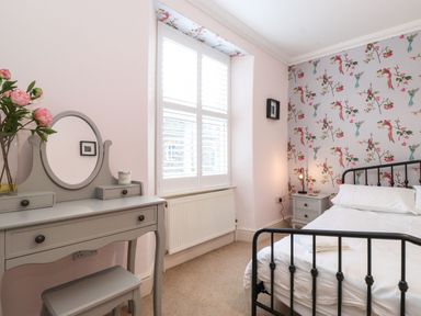 A bedroom with a bed with white bedding a grey dressing table with mirror a stool and a window with white shutters at Snowflake Cottage in Windermere