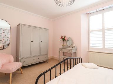 A bedroom with a gray wardrobe pink chair dressing table with flowers and a bed with white linens at Snowflake Cottage in Windermere