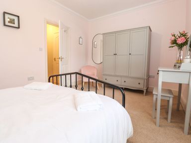 A bedroom with a black metal bed frame white bedding gray wardrobe pink chair and gray vanity table with flowers at Snowflake Cottage in Windermere