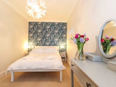 A bedroom with a bed facing a floral wallpapered wall two bedside tables and a dressing table with a mirror and a vase of flowers at Snowflake Cottage in Windermere
