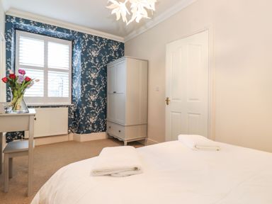 A bedroom with a bed two folded towels a gray wardrobe a desk with flowers and a window with white shutters at Snowflake Cottage in Windermere