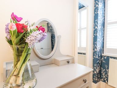 A bouquet of flowers in a glass vase on a white dressing table with a round mirror at Snowflake Cottage in Windermere