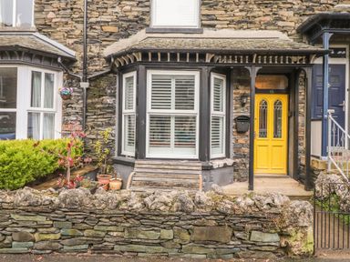 A stone house exterior with a yellow door window bench and plants behind a stone wall at Snowflake Cottage in Windermere