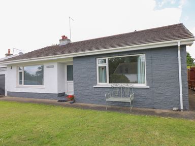 A single story house with white and gray exterior walls a green metal bench on the front patio and a grass lawn at Glynderi in Cilgerran
