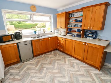 A kitchen with wooden cabinets a dishwasher microwave air fryer and open shelves with dishes by a window at Glynderi in Cilgerran