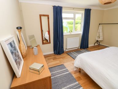 A bedroom with a wooden dresser holding a vase framed picture and mirrors next to a window with blue curtains and a bed with white bedding at Glynderi in Cilgerran