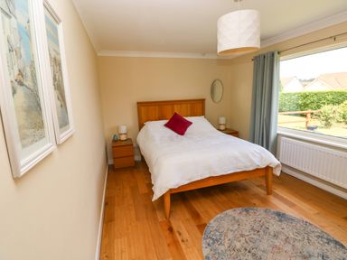 A bedroom with a wooden double bed white bedding red cushion two wooden bedside tables with lamps a large window with curtains and a circular rug on wooden floor at Glynderi in Cilgerran