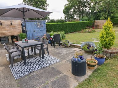 A garden patio with a table and chairs under an umbrella several potted plants a blue shed and green hedges at Glynderi in Cilgerran