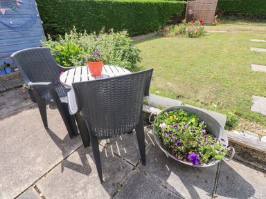 An outdoor patio area with two black chairs and a white round table with a potted plant a metal tub planted with flowers and a lawn with flowerbeds at Glynderi in Cilgerran