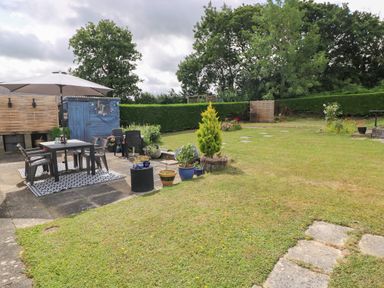 A garden with a patio area including a table and chairs under an umbrella a blue shed potted plants and a lawn with trees and hedges at Glynderi in Cilgerran