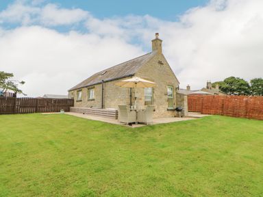 Stone house with a patio table and umbrella on a lawn with wooden fences at The Byre in Longwitton near Morpeth