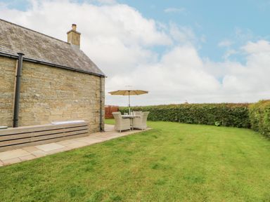 A stone building with a patio area with a table chairs and umbrella on a lawn at The Byre in Longwitton near Morpeth