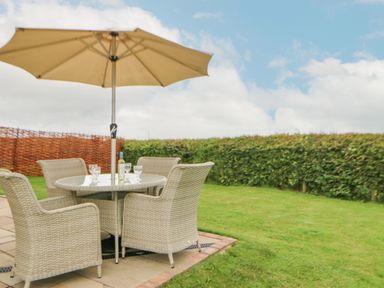 An outdoor seating area with a round table and four wicker chairs under a large umbrella on a patio with green grass and hedges surrounding at The Byre in Longwitton near Morpeth