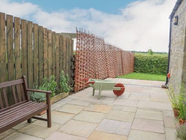A patio with a wooden bench potted plants a wooden wheelbarrow and woven fence panels at The Byre in Longwitton near Morpeth