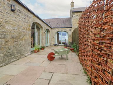 A stone patio with a wheelbarrow and potted plants near arched windows at The Byre in Longwitton near Morpeth