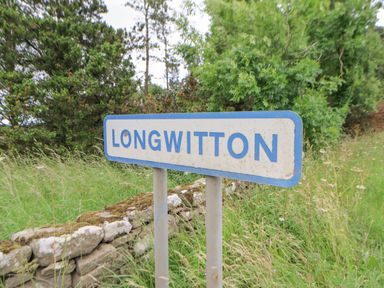 A metal sign reading Longwitton on two posts surrounded by grass trees and a stone wall in the background at The Byre in Longwitton near Morpeth