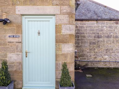 A blue front door with a brass handle and a knocker on a stone wall with two potted plants at The Byre in Longwitton near Morpeth