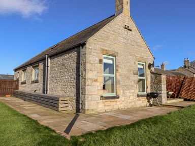 Exterior stone wall of a house with two windows a barbecue on a paved patio and grassy lawn