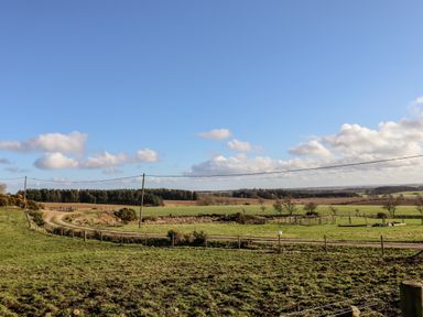 A rural landscape with green fields dirt road and wooden fences under a blue sky at The Byre in Longwitton near Morpeth
